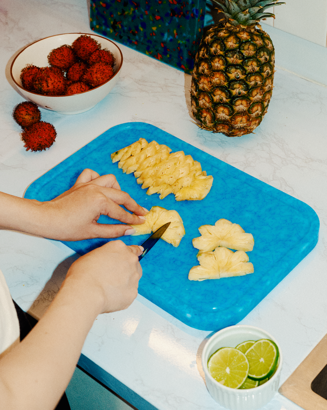 Hands slicing pineapple on a blue recycled plastic cutting board with tropical fruit and lime slices