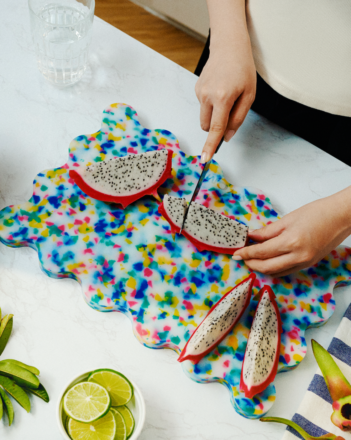 Hands slicing dragon fruit on the Chroma wavy cutting board with multicolour speckles beside limes on a kitchen countertop