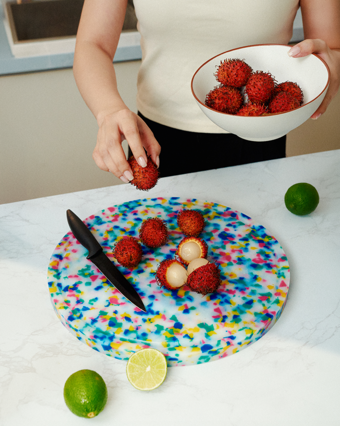 Hands placing rambutans on the Parade round cutting board with multicolour speckles beside limes on a kitchen countertop